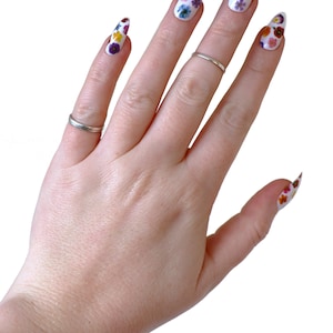 May include: A hand with long, almond-shaped nails painted white with colorful pressed flowers. The nails are decorated with a variety of flowers in shades of pink, purple, yellow, and orange.