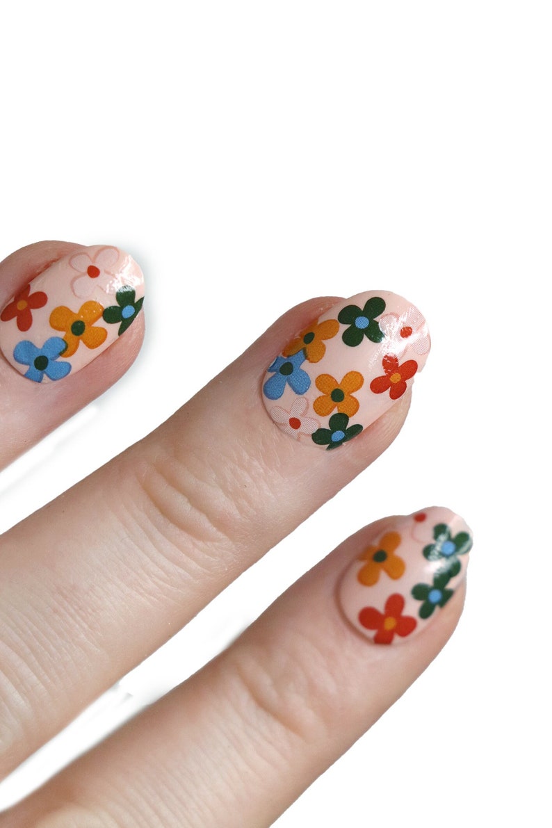K&ouml;nnte beinhalten: Nahaufnahme von drei Fingern mit hellrosa Nagellack und bunter Blumen-Nagelkunst.