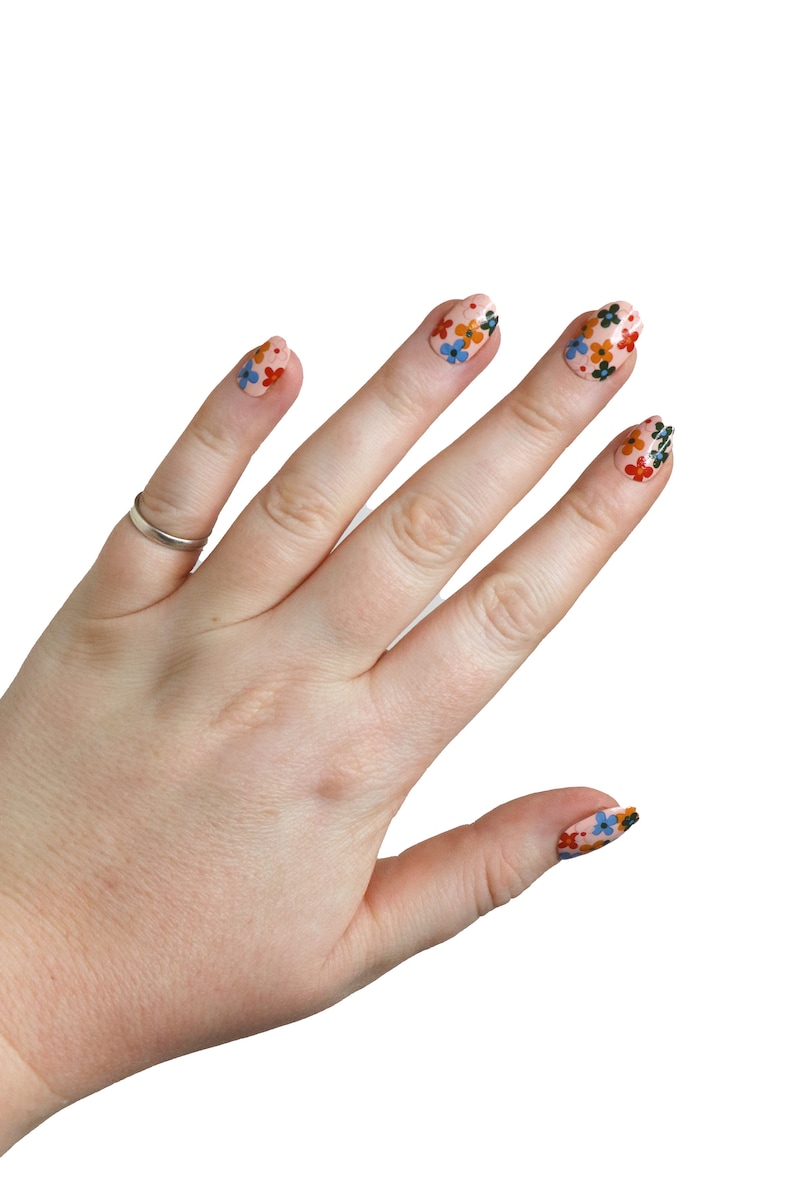 K&ouml;nnte beinhalten: Eine Hand mit rosafarbenem Nagellack und Blumenmustern auf den N&auml;geln. Die N&auml;gel sind oval geformt und die Blumen sind rot, orange, blau und gr&uuml;n.