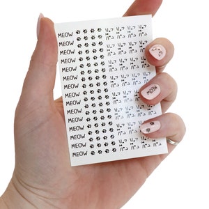 May include: A sheet of white paper with black paw prints and the word "MEOW" repeated multiple times. The paper is being held by a hand with pink nail polish and gold glitter accents.