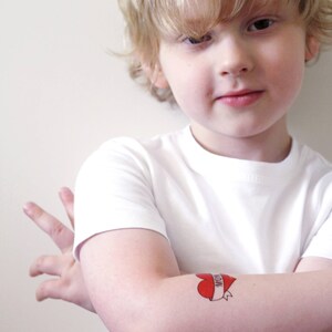 May include: A young boy wearing a white t-shirt with a red heart tattoo that says "Mom" on his arm.