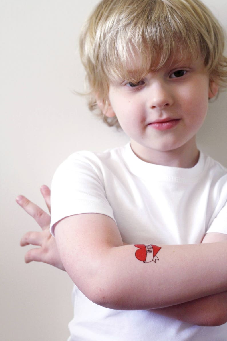 May include: A young boy wearing a white t-shirt with a red heart tattoo that says "Mom" on his arm.