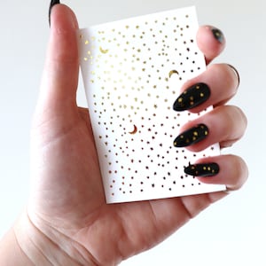 May include: A sheet of gold star and crescent moon nail decals on a white background. The decals are small and perfect for adding a touch of sparkle to your nails.