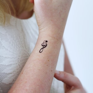 Puede incluir: Primer plano de la mu&ntilde;eca de una persona con un peque&ntilde;o tatuaje temporal negro en forma de clave de sol.