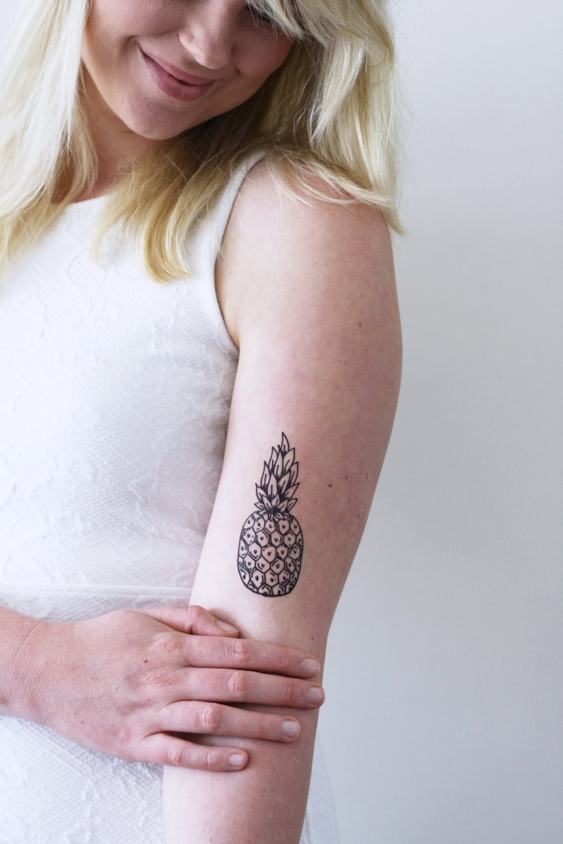 May include: A black and white line drawing of a pineapple on a person's arm. The pineapple is a simple design with a detailed outline of the fruit and leaves.
