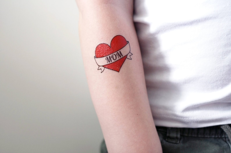 May include: A red heart with a white banner that says "MOM" on it. The heart is a temporary tattoo.