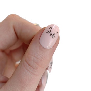 May include: A close-up of a finger with a light pink nail polish and a black cat face design.