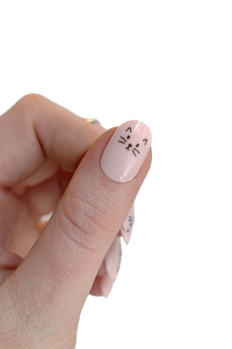 May include: A close-up of a finger with a light pink nail polish and a black cat face design.