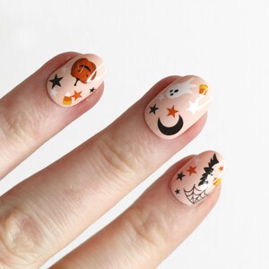 May include: Close-up of a hand with Halloween-themed nail art. The nails are painted a light pink color and decorated with black stars, orange pumpkins, white ghosts, and candy corn.