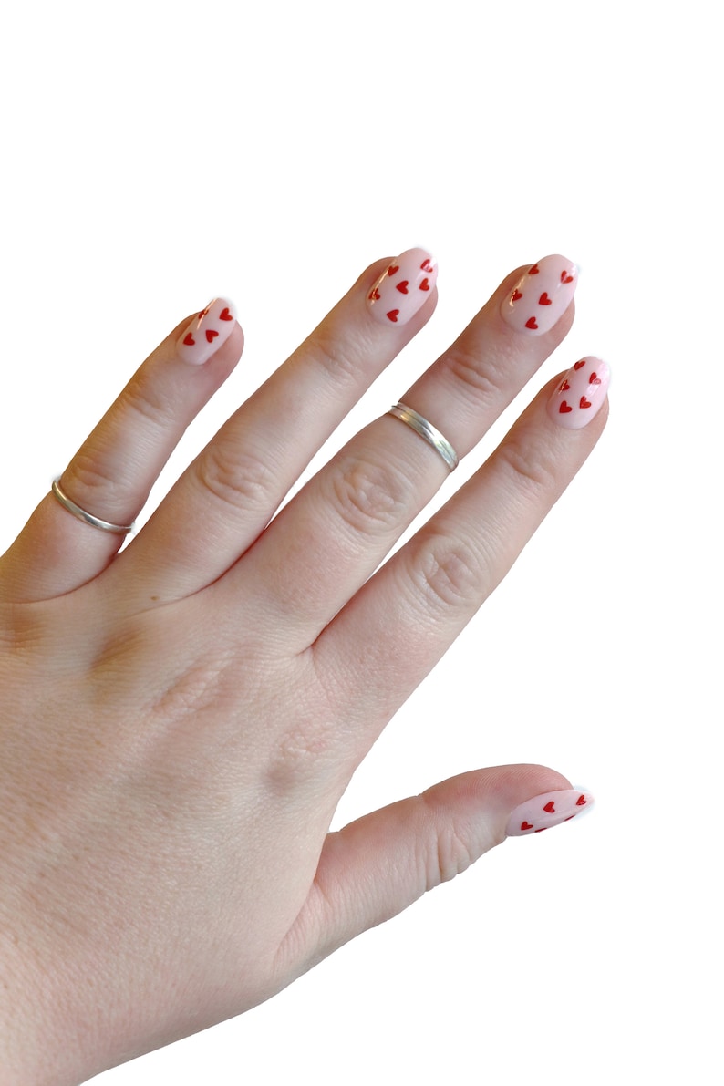 May include: A hand with pink nail polish with red heart designs on each finger.