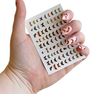 May include: A sheet of nail art stickers featuring a variety of colorful butterfly designs. The sheet is held in a hand with pink nail polish and butterfly stickers applied to the nails.