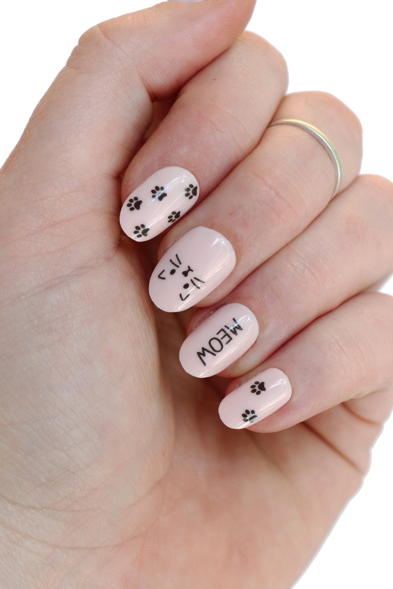 May include: A hand with pink nail polish and black cat-themed nail art. The nails have paw prints, a cat face, and the word "MEOW".