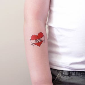 May include: A red heart with a white banner that says "MOM" on it. The heart is a temporary tattoo.