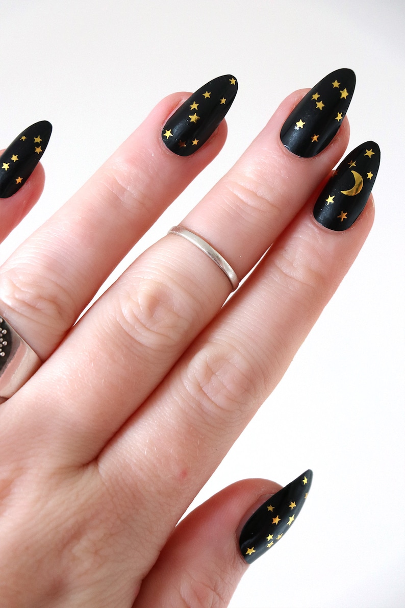 May include: Close-up of a hand with black almond-shaped nails decorated with gold stars and a crescent moon.