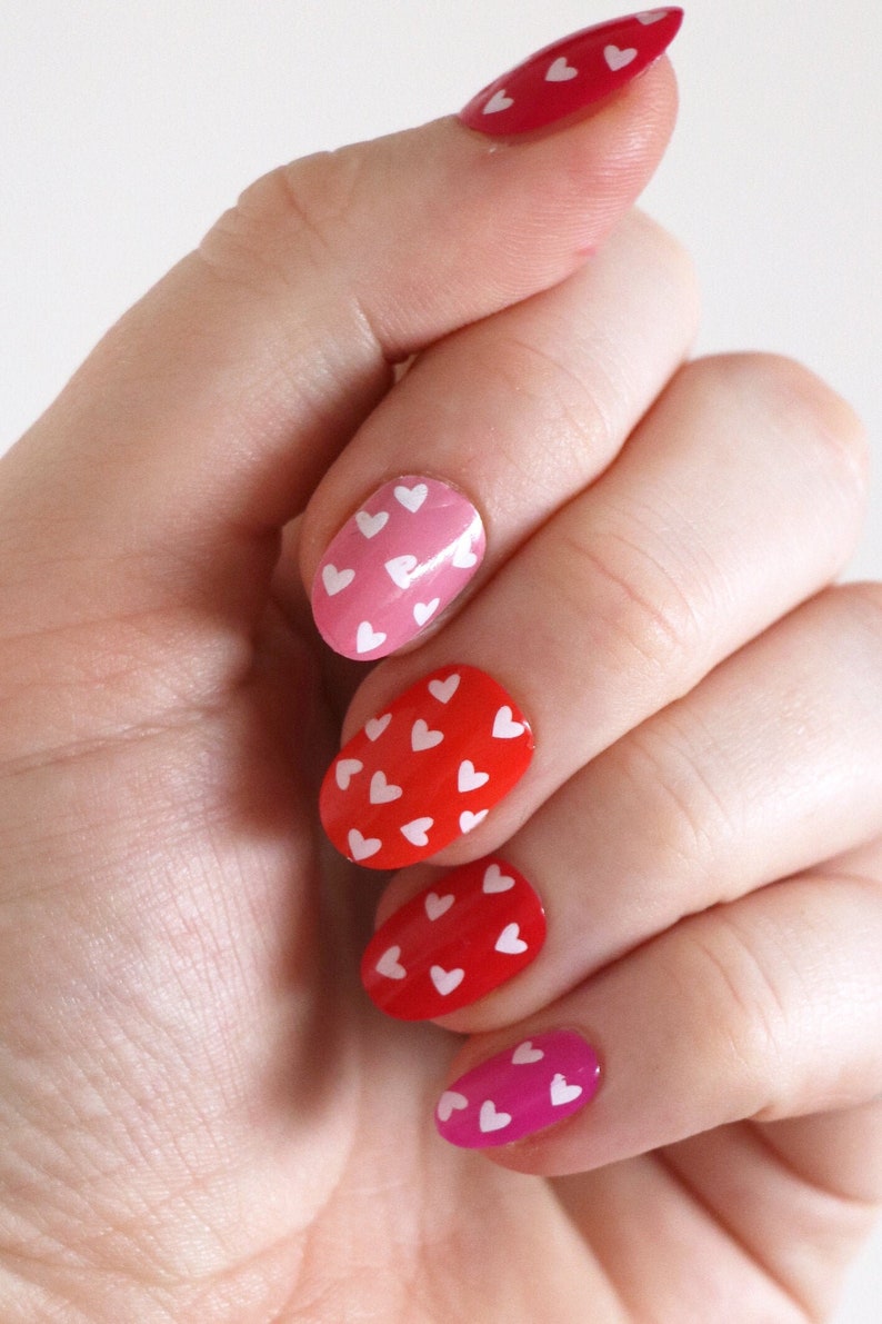 K&ouml;nnte beinhalten: Eine Hand mit f&uuml;nf Fingern&auml;geln, die in einem Farbverlauf von Rot, Rosa und Magenta lackiert sind. Jeder Nagel ist mit wei&szlig;en herzf&ouml;rmigen Aufklebern verziert.