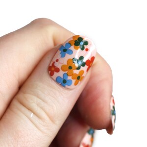 K&ouml;nnte beinhalten: Nahaufnahme eines Fingers mit einem hellrosa Nagellack-Grund und einem Blumendesign mit blauen, orangefarbenen und gr&uuml;nen Blumen.
