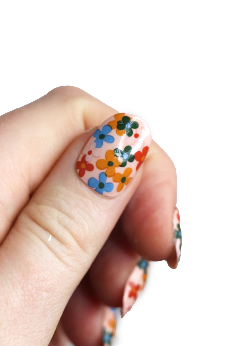K&ouml;nnte beinhalten: Nahaufnahme eines Fingers mit einem hellrosa Nagellack-Grund und einem Blumendesign mit blauen, orangefarbenen und gr&uuml;nen Blumen.