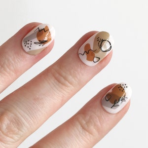 May include: A hand model wearing oval-shaped press-on nails with abstract designs in brown, beige, and white. The nails feature simple line drawings and geometric shapes.