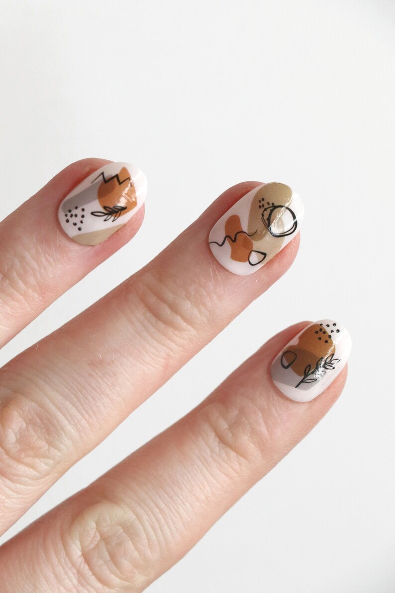 May include: A hand model wearing oval-shaped press-on nails with abstract designs in brown, beige, and white. The nails feature simple line drawings and geometric shapes.