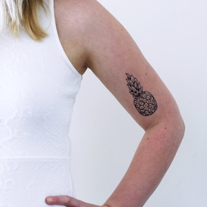 May include: A woman's arm with a black and white pineapple tattoo on the upper arm.