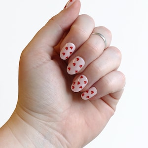 May include: A hand with oval-shaped nails painted a light pink color with red heart designs.