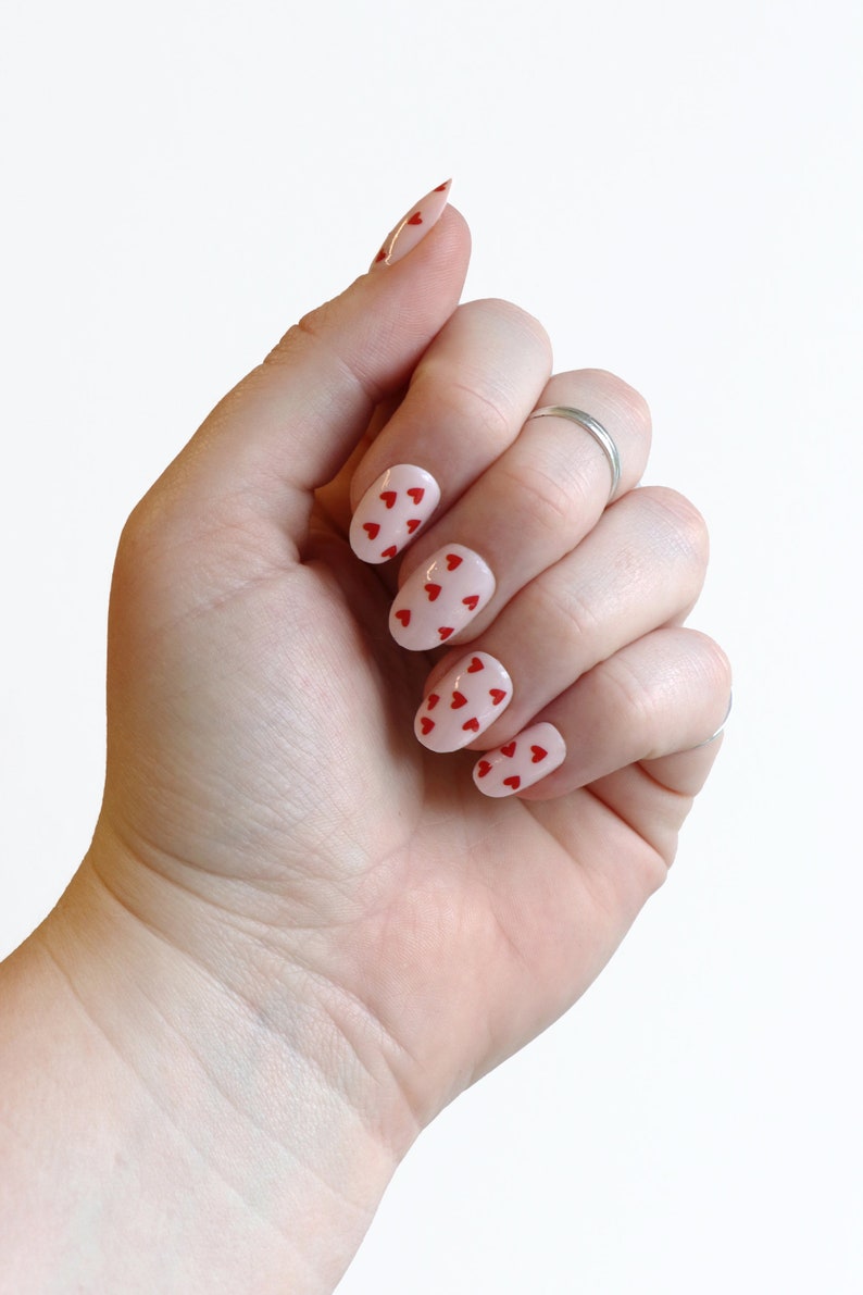 May include: A hand with oval-shaped nails painted a light pink color with red heart designs.