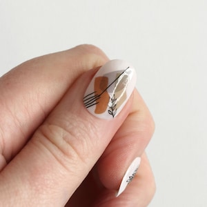May include: Close-up of a finger with a white oval-shaped nail with an abstract design featuring brown and black lines, a brown shape, and a small white branch with leaves.