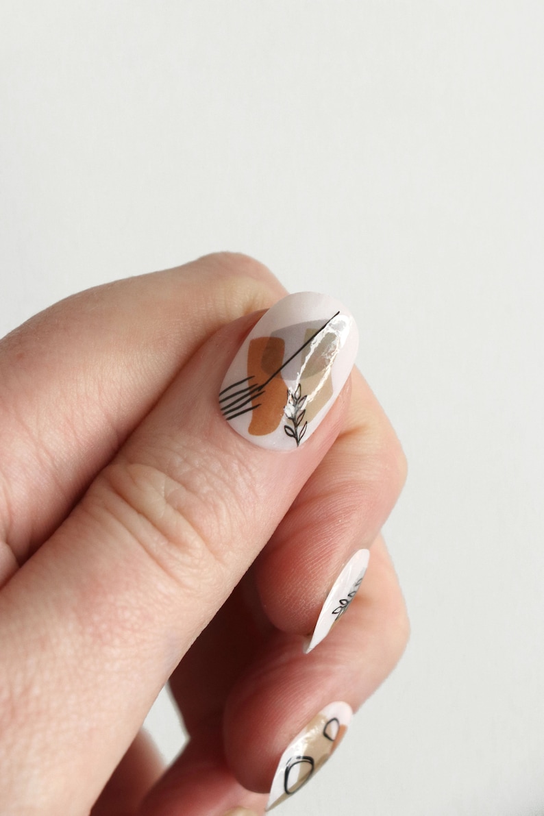 May include: Close-up of a finger with a white oval-shaped nail with an abstract design featuring brown and black lines, a brown shape, and a small white branch with leaves.