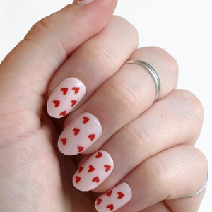 May include: A hand with oval-shaped nails painted a light pink color with small red hearts on each nail.