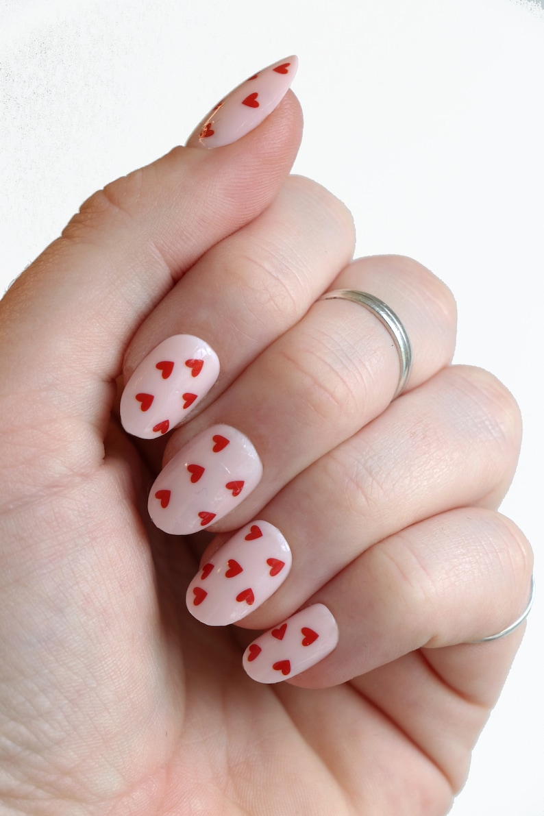 May include: A hand with oval-shaped nails painted a light pink color with small red hearts on each nail.