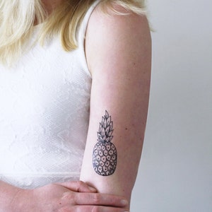 May include: A black and white line drawing of a pineapple on a person's arm.