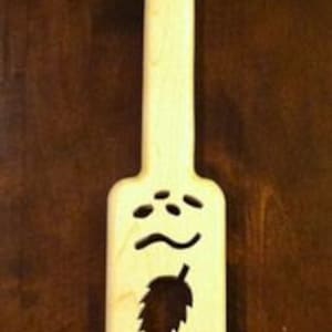 May include: A wooden paddle with a carved design of a hop leaf and a wheat stalk. The paddle is light brown and has a smooth finish.