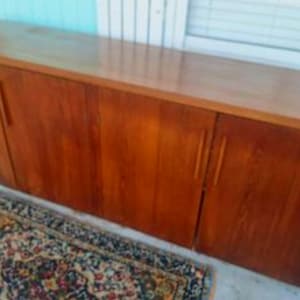 May include: A long, wooden cabinet with four doors. The cabinet is a warm, medium brown color with a smooth, glossy finish. The doors have vertical, rectangular handles. A patterned rug is in the foreground.
