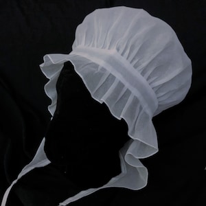 May include: A white, ruffled, sheer bonnet with a gathered crown. The bonnet is made of a lightweight fabric and has a simple, elegant design.