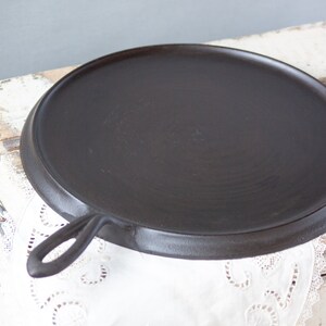 May include: A black cast iron griddle with a single handle. The griddle is sitting on a white surface with a lace doily.