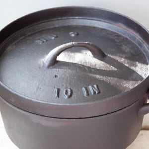 May include: A dark gray cast iron Dutch oven with a matching lid. The lid has a handle and the text "10 IN" embossed on it. The pot has a handle on the side. This cookware is suitable for outdoor cooking.