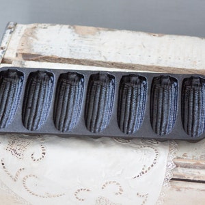 May include: A black cast iron cornbread pan with six individual molds shaped like corn cobs. The pan is sitting on a white wooden surface.