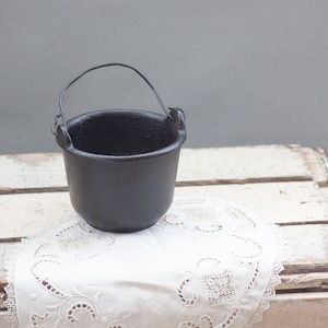 May include: A small black cast iron pot with a wire handle. The pot is sitting on a white doily on a weathered white wooden crate.