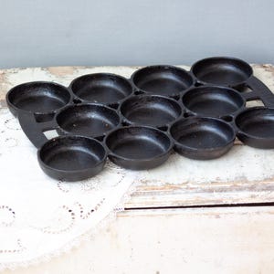 May include: Black cast iron muffin pan with 12 individual round molds. The pan is on a white lace doily and a distressed white wooden surface. The pan is designed for baking muffins or small cakes.