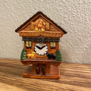 May include: A miniature cuckoo clock with a brown wooden-style design. The clock features a white clock face, small windows, and a decorative owl on top. A yellow deer and black bear are on the clock's shelf.
