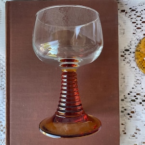 May include: A clear glass wine goblet with a brown, ribbed stem and base. The goblet is sitting on a brown book. The glass has a rounded bowl and is designed for wine.