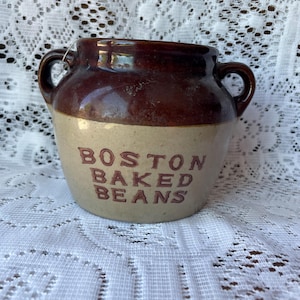 May include: A vintage, two-toned ceramic crock with a brown top and tan base. The words "BOSTON BAKED BEANS" are printed in red on the front. The crock has two small handles and a glossy finish, perfect for kitchen decor.