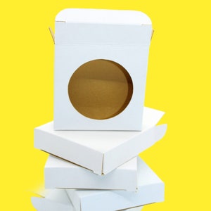 Può includere: Una pila di scatole di cartone bianco di forma rettangolare. La scatola superiore presenta una finestra circolare ritagliata sul pannello frontale, che rivela l'interno marrone della scatola. Le scatole sono realizzate in cartone bianco opaco.