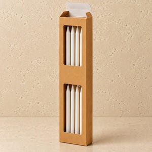 May include: A tall, rectangular brown cardboard box with two vertical cut-out windows displaying eight slender, white taper candles. The box is standing upright, featuring a top flap that is partially open, revealing the white candles inside.