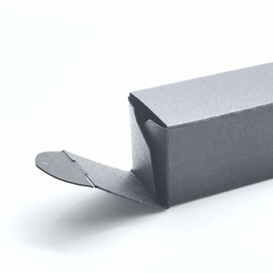 May include: A rectangular black cardboard box with a partially opened flap. The box features a smooth matte finish and a distinct folding mechanism on the side, showing the construction of the paper packaging against a plain white background.