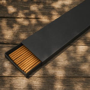 May include: A black rectangular box, partially open, revealing a collection of incense sticks. The incense sticks are a light brown color and are neatly arranged inside the box. The box is set on a wooden surface with natural light and shadows.