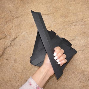 Puede incluir: Una mano sosteniendo una pila de papel negro doblado. El papel está dispuesto en una forma geométrica, con piezas largas y rectangulares que se extienden hacia arriba. El fondo es una superficie texturizada de color beige.