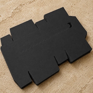 May include: A stack of unfolded black cardboard boxes with cut-out tabs and a handle. The boxes are neatly arranged on a light brown, textured surface. The boxes are designed to be folded into a container.
