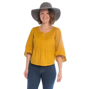 Könnte beinhalten: Eine Frau trägt eine senfgelbe Bluse mit weitem Ausschnitt und Glockenärmeln. Sie trägt auch blaue Jeans und einen grauen Fedora.