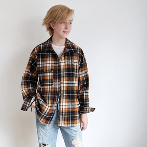 May include: A person wearing a brown and black plaid flannel shirt with a white undershirt, blue jeans with rips, and black sneakers.